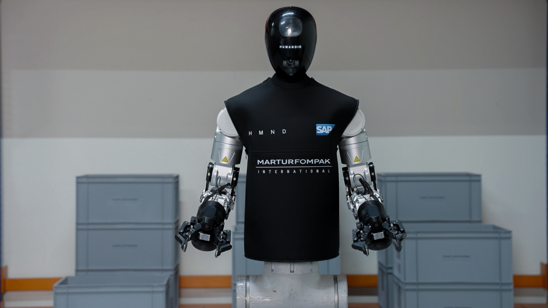 The HMND 01 Alpha humanoid robot standing in an industrial facility in a black vest branded HMND, SAP and Martur Fompak, with two arms ending in parallel grippers and grey logistics totes stacked around it.